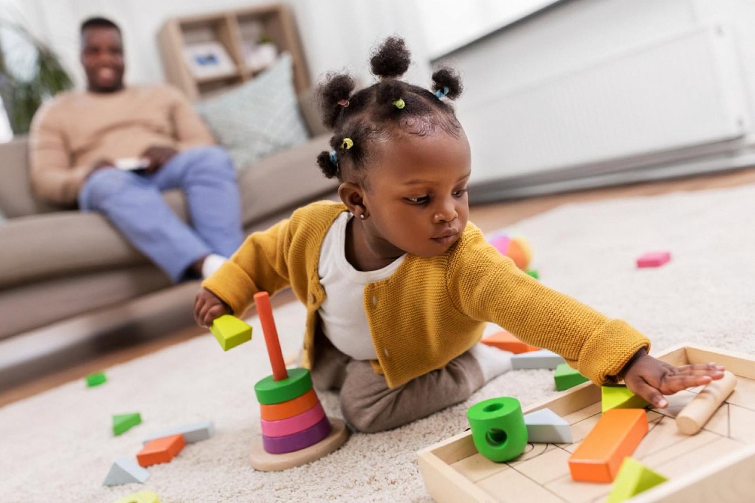 Preschool girl arranging wooden Montessori blocks while her father watches from the sofa
